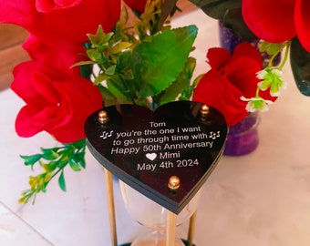 Op de afbeelding: Zwarte hartvormige zandloper met gouden accenten en een gepersonaliseerd bericht: "Tom, jij bent degene met wie ik door de tijd wil gaan. Gefeliciteerd met jullie 50e huwelijksverjaardag Mimi 4 mei 2024".