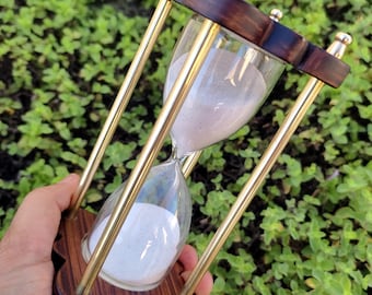 Könnte beinhalten: Eine Sanduhr aus Holz mit Messingakzenten und einem weißen Sandtimer. Die Sanduhr wird in einer Hand vor einem grünen, belaubten Hintergrund gehalten.