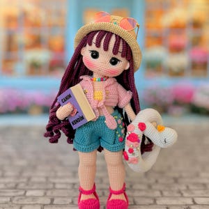 May include: A crocheted doll with long dark purple hair, wearing a beige straw hat with orange sunglasses, a pink cropped cardigan, a yellow top, and teal shorts. The doll holds a book titled CANIM KENDIM and a white floral-decorated swim ring.