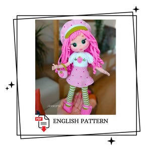 Op de afbeelding: Een handgemaakte gehaakte pop met roze haar, een bijpassende baret en een roze jurk. De pop houdt een kleine handtas vast en draagt gestreepte groen-witte sokken. Het shirt van de pop heeft een aardbeienontwerp. De tekst "ENGLISH PATTERN" is zichtbaar.