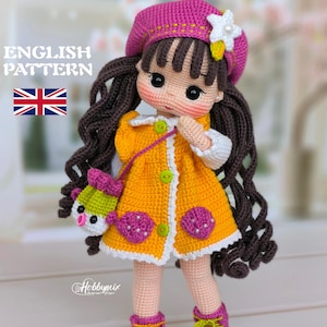 Puede incluir: Muñeca de ganchillo con vestido amarillo, boina rosa y botas a juego. La muñeca tiene pelo largo castaño y un pequeño bolso. El texto "ENGLISH PATTERN" y una bandera británica son visibles.