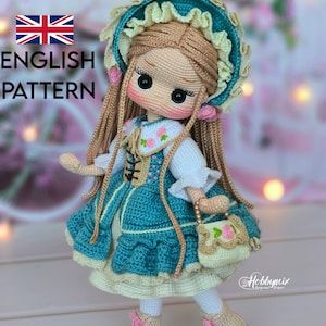 Pode incluir: Uma boneca de crochê feita à mão, com cabelo castanho comprido, vestida com um vestido azul com detalhes brancos, um chapéu a condizer e botas bege com atacadores cor-de-rosa. A boneca segura uma pequena bolsa com um desenho floral. O texto "ENGLISH PATTERN" é visível.