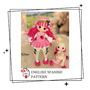 May include: A handmade crochet doll with pink hair, a pink dress, and a matching hat. The doll has a strawberry detail on the dress. A small crochet bear is also present. The image includes the text "ENGLISH SPANISH PATTERN".