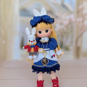 Könnte beinhalten: Eine gehäkelte Puppe mit langem blonden Haar, bekleidet mit einem blauen Kleid mit weißer Schürze, einer blauen Haarschleife und roten Schuhen. Die Puppe hält eine kleine weiße Hasenfigur mit roter Jacke und blauer Fliege. Eine goldfarbene Halskette mit Uhrenanhänger hängt um den Hals der Puppe, und ein herzförmiges Emblem ist an der Schürze befestigt.