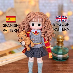 Può includere: Bambola all'uncinetto fatta a mano con capelli ricci castani, che indossa un maglione grigio, una gonna a pieghe e una sciarpa rossa e gialla. La bambola tiene in mano un piccolo orsacchiotto. L'immagine include il testo "Spanish Pattern" e "English Pattern".