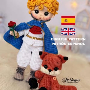 Puede incluir: Muñeco de ganchillo de un joven príncipe con capa azul, sosteniendo una rosa roja, y un zorro de ganchillo. El príncipe tiene cabello rizado dorado y una corona dorada. El zorro es naranja y blanco. Incluye el texto "ENGLISH PATTERN PATRÓN ESPAÑOL".