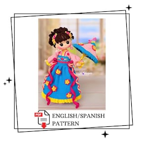 Pode incluir: Uma boneca de crochê com cabelo castanho escuro, vestindo um top rosa e uma saia azul decorada com flores amarelas e rosas. A boneca segura um pequeno guarda-chuva rosa e azul com detalhes amarelos. O texto "Hobbymix" e "ENGLISH/SPANISH PATTERN" aparecem.