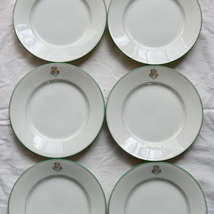 Peut inclure: Six assiettes en céramique blanche avec un bord vert et un motif de crête dorée. Les assiettes sont disposées en deux rangées de trois sur une surface blanche. Les assiettes sont rondes et ont un design simple et élégant.