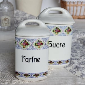 Puede incluir: Dos botes de cerámica blanca con tapas, etiquetados "Farine" y "Sucre" en escritura negra. Cada bote presenta un diseño floral con un borde azul y verde. Los botes tienen un asa en la parte superior.
