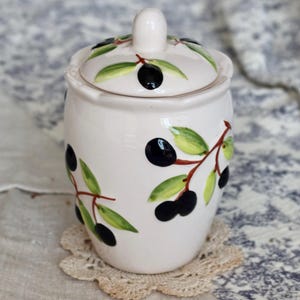 Peut inclure: Un pot en céramique blanc avec un couvercle, décoré de motifs de branches d'olives noires et de feuilles vertes. Le pot est posé sur un petit napperon rond en dentelle beige. L'arrière-plan comprend un tissu à motifs.