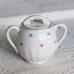 May include: White ceramic sugar bowl with a lid and two handles. The bowl is decorated with small pink floral patterns and gold trim. The sugar bowl is sitting on a white lace doily.