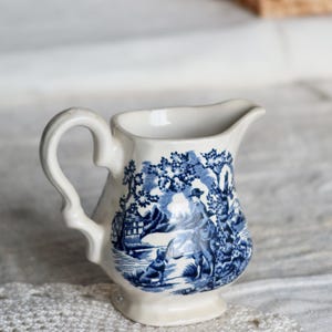Puede incluir: Jarra pequeña de cerámica blanca con una escena pastoral azul y blanca. La jarra tiene un asa curva y un pico para verter. El diseño presenta figuras, árboles y edificios. Una pieza decorativa para servir.
