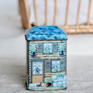 May include: A decorative metal tin shaped like a house, featuring a blue tiled roof and a detailed facade with windows, shutters, and a door. The tin has a rustic, vintage aesthetic, with a small dog illustration near the door.