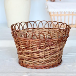 Può includere: Un cestino marrone intrecciato con un bordo smerlato. Il cestino è realizzato in vimini naturale e ha una forma arrotondata. Viene probabilmente utilizzato per la conservazione o l'esposizione, con un diametro di circa 20 cm.