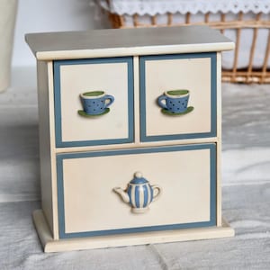 Peut inclure: Petit meuble décoratif en bois à trois tiroirs. Le meuble est peint en crème avec des bordures bleues. Les deux tiroirs supérieurs présentent des tasses à thé bleues avec des rebords verts et des soucoupes. Le tiroir inférieur a une théière bleue et blanche.