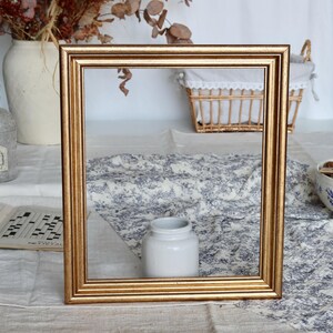 Peut inclure: Un cadre photo carré de couleur dorée. Le cadre est vide, avec un design simple et élégant. L'arrière-plan comprend un vase blanc avec des fleurs séchées, un panier et une nappe à motifs.