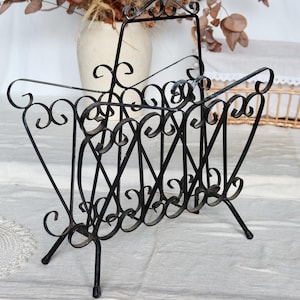 Può includere: Portariviste in ferro battuto nero con un design a volute decorative. Il portariviste ha una maniglia e tre gambe. Un vaso con fiori secchi è sullo sfondo.