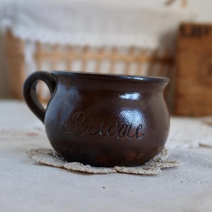 以下が含まれることがあります： ハンドルが付いた茶色の陶器製のマグカップで、側面に「Beurre」と刻まれています。マグカップは白いレースのコースターの上に置かれています。