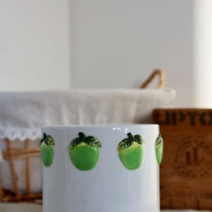 May include: White ceramic mug with green apple accents around the rim. The mug is sitting on a white crocheted doily.