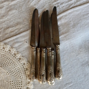 Peut inclure: Ensemble de couteaux anciens en argent avec des manches ornés. Les lames présentent une légère patine, et les manches sont décorés de motifs floraux et ovales détaillés. Les couteaux sont disposés sur une surface texturée blanc cassé, évoquant le vintage.
