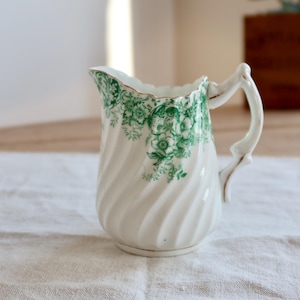 Peut inclure: Pichet en céramique blanche avec un motif floral vert. Le pichet a un corps torsadé et une anse incurvée. Le bord est festonné et orné d'or. Parfait pour servir des boissons ou comme vase décoratif.