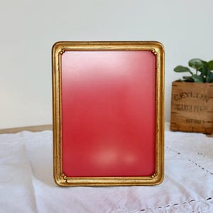 Könnte beinhalten: Ein rechteckiger goldfarbener Bilderrahmen mit rotem Innenraum. Der Rahmen hat ein detailliertes, verziertes Design. Der Rahmen steht auf einer weißen Spitzendecke. Eine kleine Holzkiste mit Text und eine Schüssel befinden sich im Hintergrund.