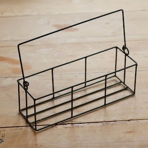 Op de afbeelding: Een zwarte metalen draadmand met een handvat. De rechthoekige mand heeft een rasterachtig ontwerp en staat op een lichtgekleurd houten oppervlak. De handgreep staat rechtop.