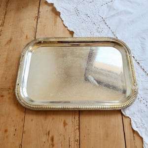 May include: A rectangular silver-toned serving tray featuring a polished, reflective surface with an intricate, subtle floral engraving in the center and a decorative beaded border along the raised edges of the metal frame.