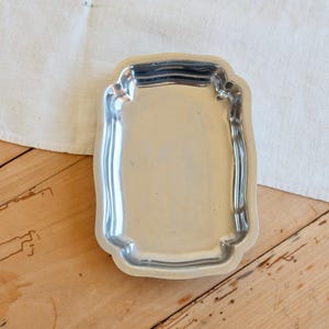 May include: A small, rectangular silver tray with a decorative, scalloped edge. The empty tray reflects light. It rests on a wooden surface, beside a white cloth. The tray is approximately 15 cm long.