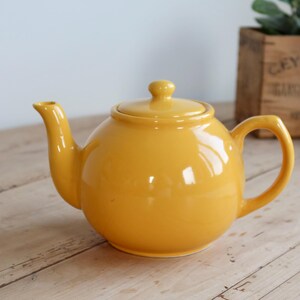 May include: A bright yellow ceramic teapot with a rounded body, a curved spout, a matching lid with a small knob handle, and a sturdy side handle. The teapot features a smooth, glossy finish and a classic, simple design for serving hot beverages.