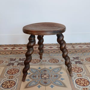 May include: A dark wooden stool with a circular seat and three spiral-turned legs. The stool is made of dark-stained wood and stands on a patterned tile floor. The design is reminiscent of antique furniture.
