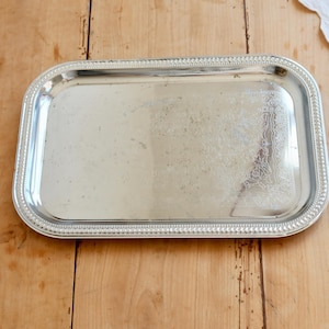 Könnte beinhalten: Ein rechteckiges silberfarbenes Serviertablett mit abgerundeten Ecken und einem dekorativen Perlenrand. Die Mitte des Tabletts weist eine glatte, reflektierende Oberfläche mit leichten Gebrauchsspuren und einem floralen Gravurmuster auf.