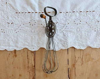 Può includere: Frusta per uova in metallo vintage con manico in legno. La frusta ha un manico circolare e un meccanismo a ingranaggi. La frusta è realizzata con sottili fili di metallo. La frusta è appoggiata su un tessuto bianco ricamato e una superficie in legno.