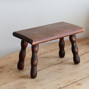 May include: A small, antique wooden step stool with a rectangular top and four curved legs. The stool is made of dark brown wood and sits on a light brown wooden surface. The stool is a vintage piece.