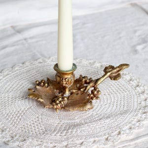 May include: A gold-colored metal candle holder with a leaf and grape design. The candle holder has a detailed, ornate design and holds a white candle. The candle holder is placed on a white, crocheted doily.