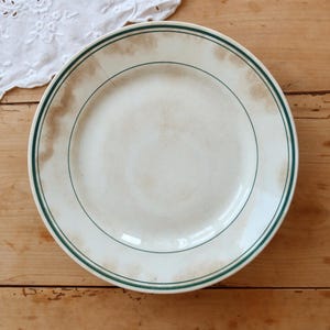 May include: A round ceramic plate featuring an off-white base with brown staining and two thin dark green circular bands along the rim. The plate shows a visible thin crack extending from the edge toward the center of the surface.