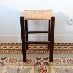 Puede incluir: Un taburete cuadrado con estructura de madera de color marrón oscuro y asiento tejido de paja en tono beige claro. El taburete cuenta con cuatro patas rectas unidas por barras de soporte horizontales en todos sus lados.