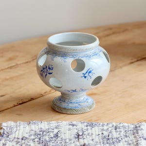 May include: A white ceramic bulbous vase featuring a series of circular cutouts around the midsection. The surface is decorated with blue floral patterns and intricate line work, finished with a textured base and a rimmed opening at the top.