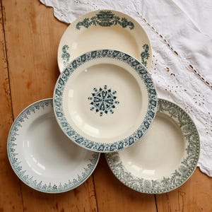 May include: A set of three round ceramic plates featuring intricate dark blue and green transferware patterns. The plates display detailed floral and geometric borders, with one plate showcasing a central star-like motif on a cream-colored base.