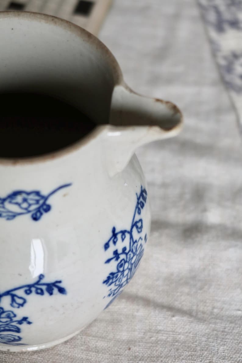 Puede incluir: Una jarra de cer&aacute;mica blanca con dise&ntilde;os florales azules. La jarra tiene un pico y un asa. El interior es oscuro. La jarra est&aacute; sobre una superficie de color claro.