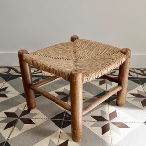 May include: A low stool with four cylindrical wooden legs connected by horizontal stretchers. The square seat is constructed from woven natural fiber cord in a light tan color, featuring a distinct diagonal pattern meeting at the center.