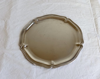Puede incluir: Una bandeja redonda de metal plateado con una superficie lisa y reflectante, y un borde decorativo festoneado con sutiles hendiduras a lo largo del perímetro. La bandeja tiene un borde elevado y una zona central plana y circular.