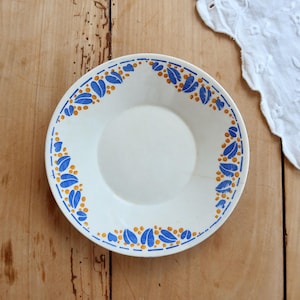 May include: A round white ceramic saucer featuring a decorative border pattern of small blue leaves and yellow dots. The design is arranged in a circular motif along the rim of the dish, which has a smooth, plain center and a slightly raised edge.