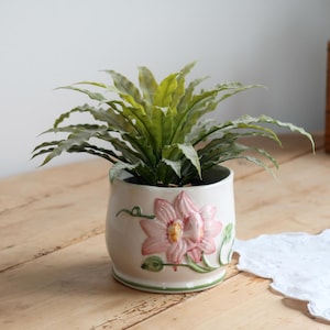Peut inclure: Un cache-pot en céramique blanche orné d'un motif en relief représentant une fleur de lotus rose avec des tiges et des feuilles vertes. Le pot contient une plante verte luxuriante aux feuilles longues, pointues et aux bords ondulés.