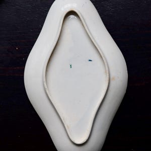 May include: A white, porcelain dish with a unique, elongated diamond shape. The dish has a raised rim and a smooth, glossy surface. The interior features subtle blue and green markings. The dish is likely vintage and suitable for serving small portions.