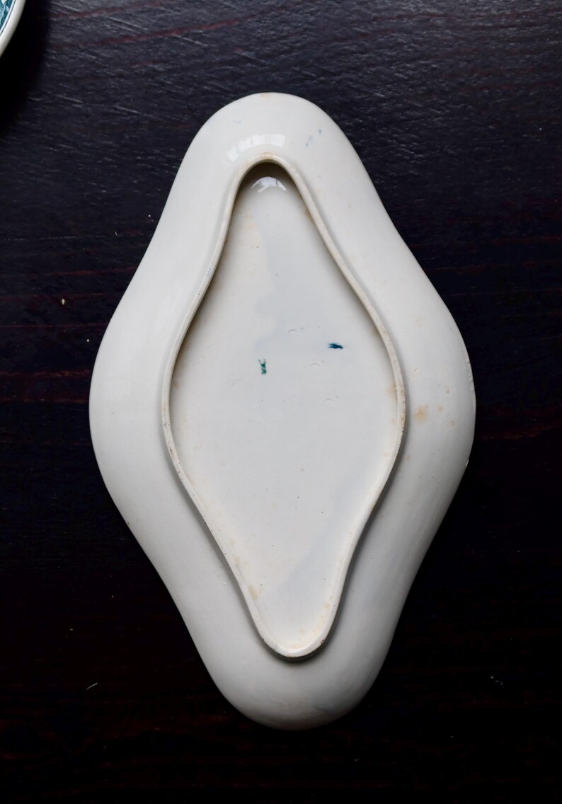 May include: A white, porcelain dish with a unique, elongated diamond shape. The dish has a raised rim and a smooth, glossy surface. The interior features subtle blue and green markings. The dish is likely vintage and suitable for serving small portions.