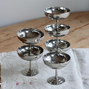 May include: A collection of silver-colored champagne coupe glasses. The glasses are arranged on a white cloth and a wooden surface. The glasses have a classic design with a round bowl and a long stem. The image shows six glasses in total.