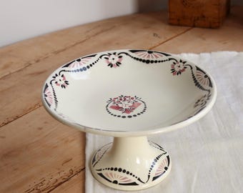 May include: A cream-coloured ceramic pedestal bowl featuring a central floral motif and a decorative border of black dots and pink fan-shaped patterns along the rim and base. The bowl has a wide, shallow basin and a sturdy, flared pedestal foot.