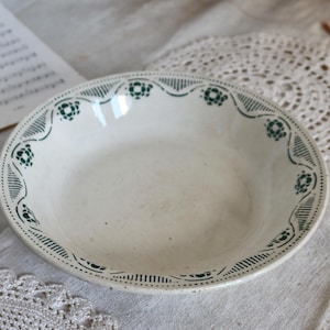 May include: A white ceramic bowl with a green floral and geometric border design. The bowl has a slightly oval shape and a shallow depth. The rim features a repeating pattern of stylized flowers and lines. The bowl is likely vintage or antique.