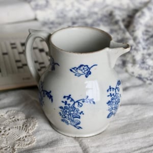 Puede incluir: Una jarra de cer&aacute;mica blanca con dise&ntilde;os florales azules. La jarra tiene un asa y un pico. El dise&ntilde;o floral se repite alrededor del cuerpo de la jarra. La jarra est&aacute; sobre una superficie blanca.